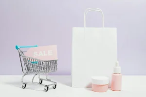 A small shopping cart holding a pink “SALE” sign beside beauty products and a white paper bag, representing branding and promotion in ecommerce.