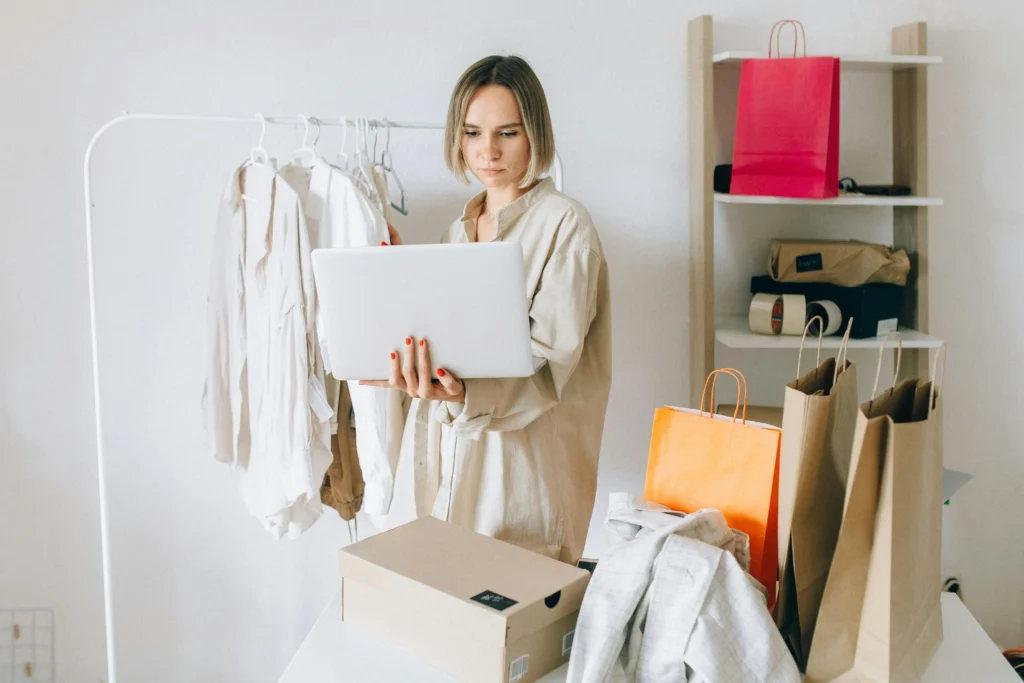 A woman standing in a minimalistic room with shopping bags and clothing, using a laptop to manage her online ecommerce store.